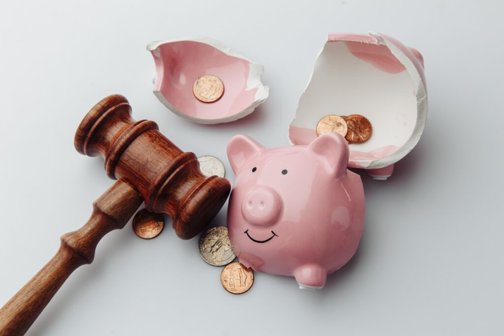 Broken piggy bank with dollar bills, coins and wooden gavel on a white table.